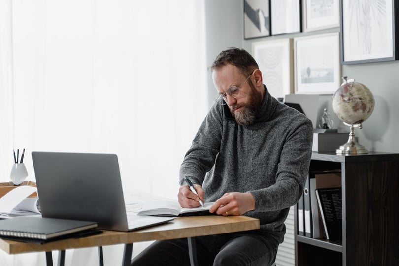 Man writing in a notebook while on phone and laptop